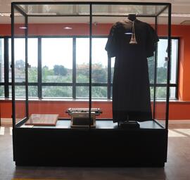 Fotografia da vitrine expositora que contém a placa, a máquina de escrever e a beca